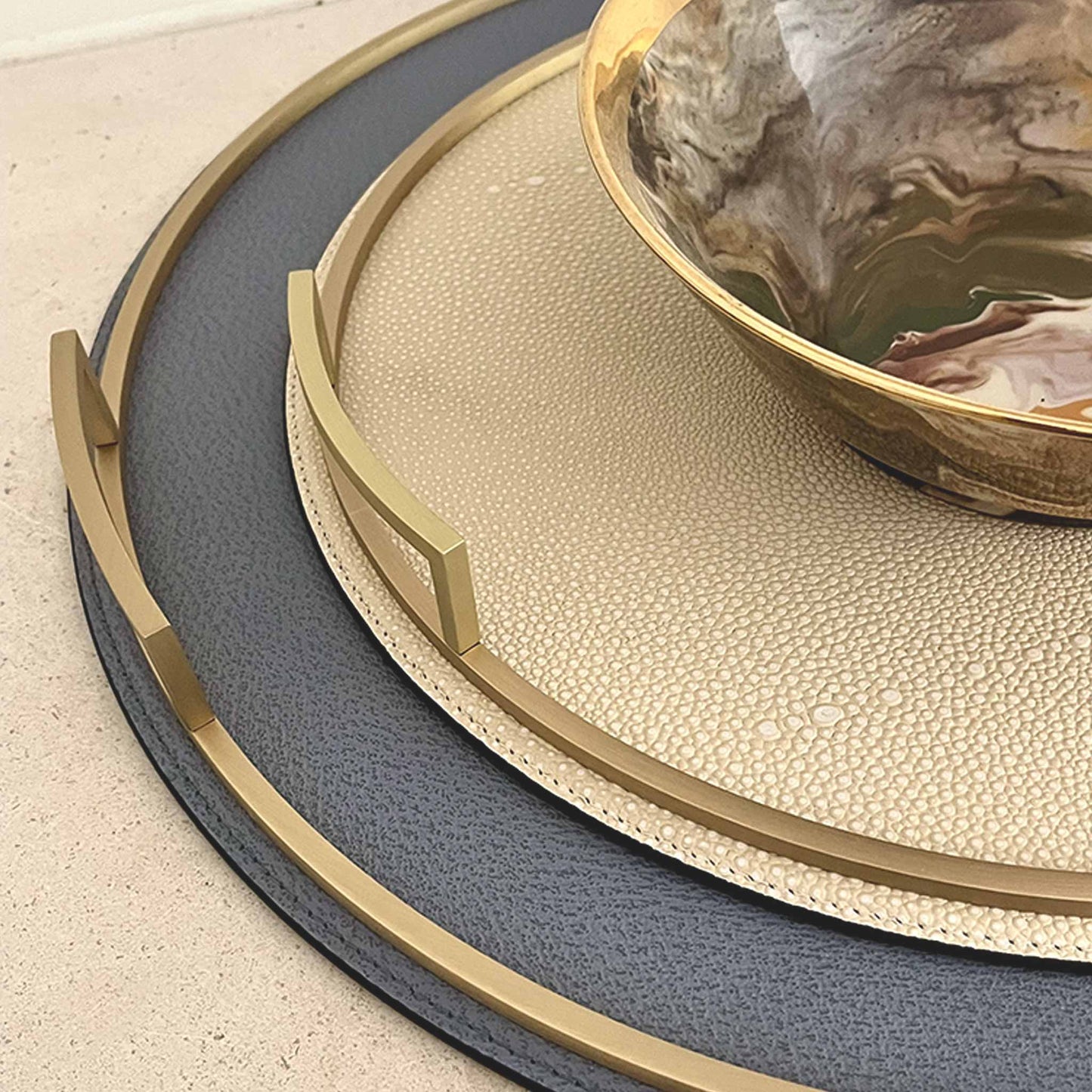 Lifestyle image with grey-blue round leather tray with smaller cream tray with shagreen texture, both with brass handles