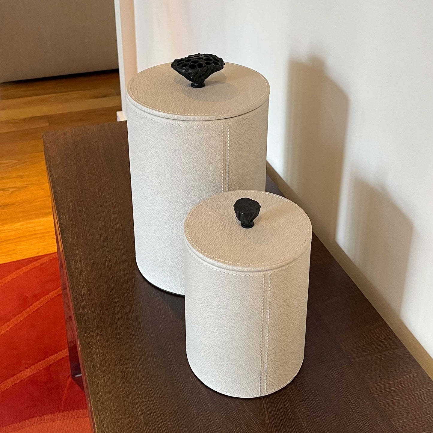 Lifestyle image of matching Close up lifestyle image of two light cream leather cylinder boxes with cast brass handles. Displayed on a luxury console