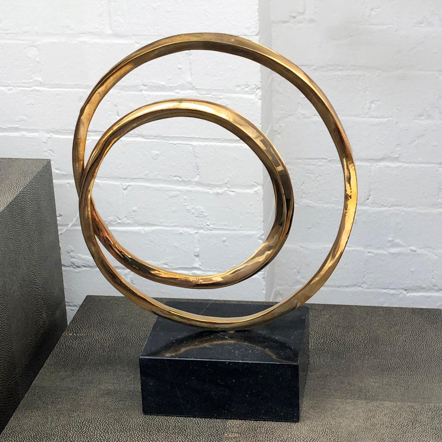 Lifestyle image of luxury round bronze decor Infinity Sculpture with marble block base, displayed on shagreen tables against a white brick wall.