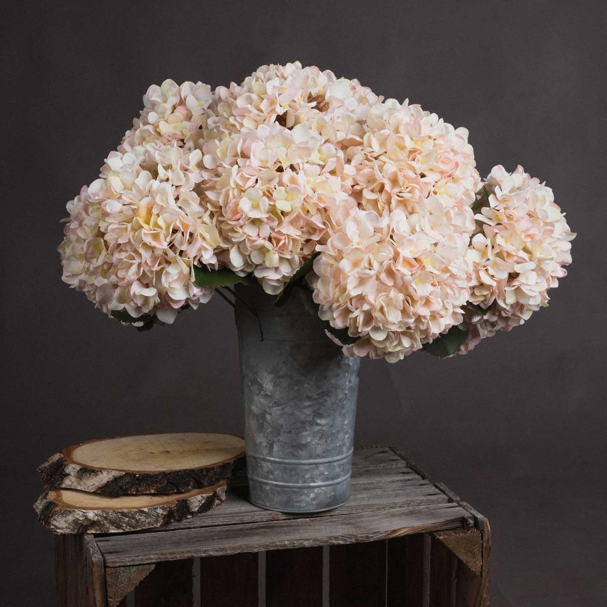 Lifestyle image of a bunch of creamy ivory and peach toned white faux hydrangea stems in a rustic bucket