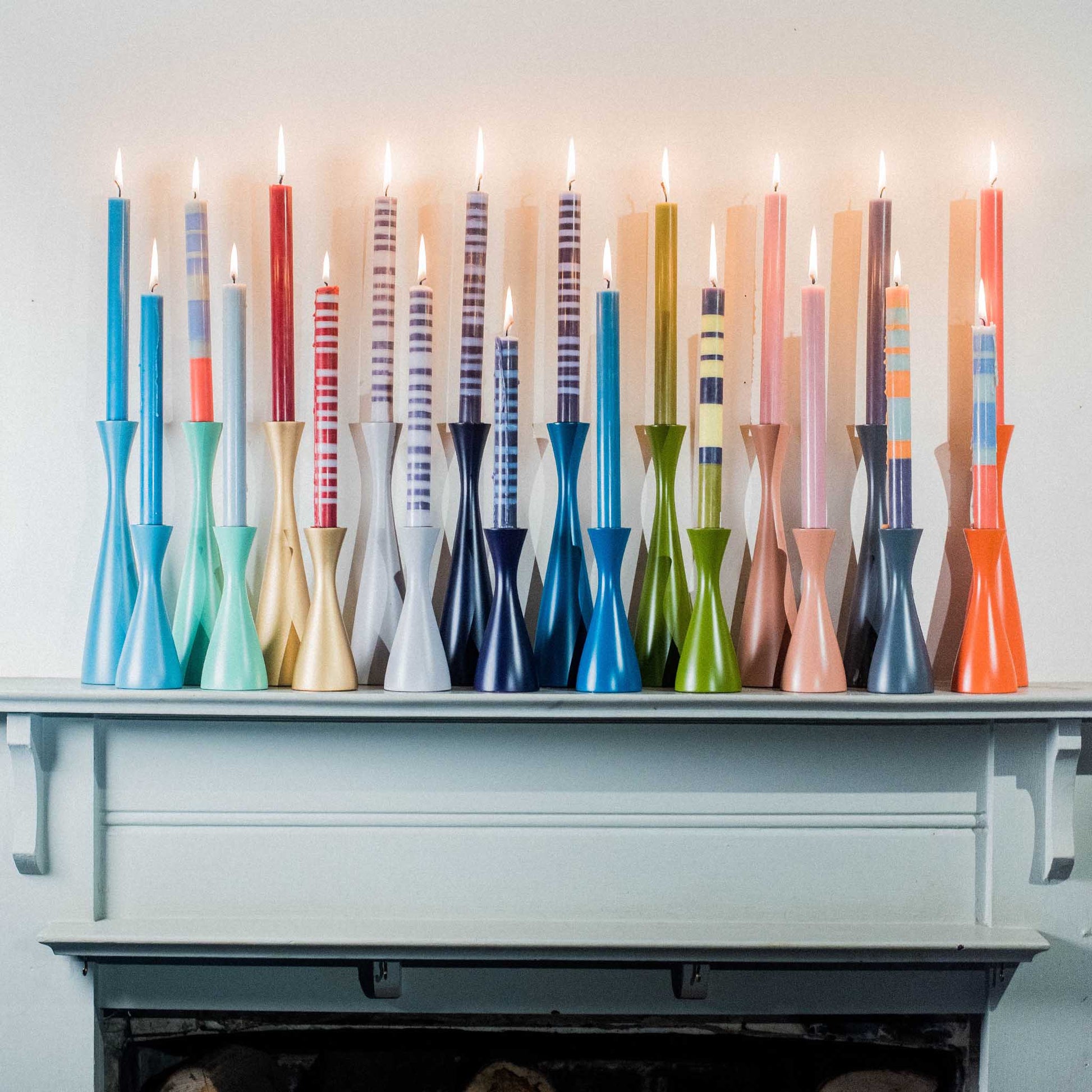 A lifestyle image of multi coloured, striped and plain tall candle collection and candle holders, placed on a fireplace mantle