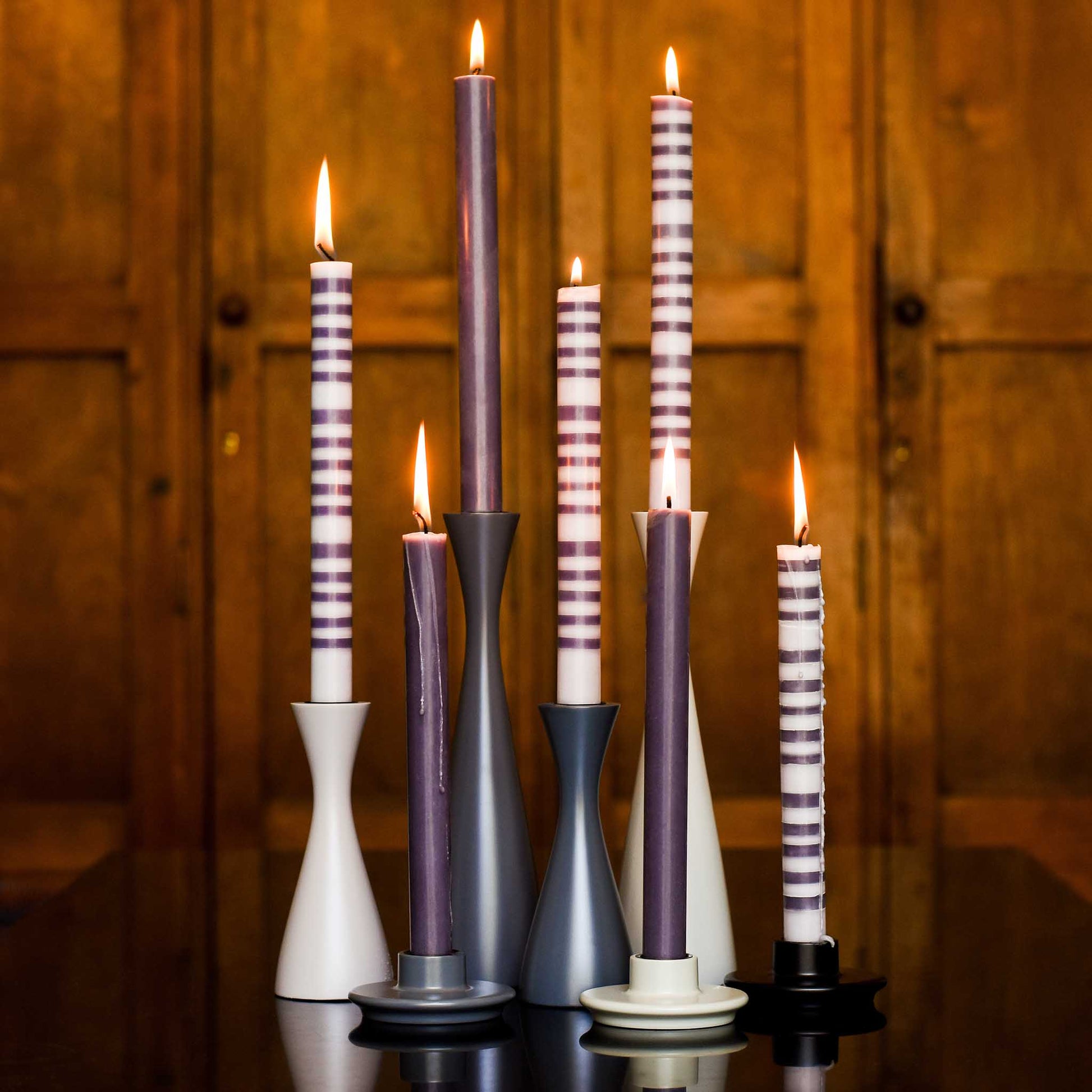 Lifestyle image of several grey and white candleholders in a variety of heights with striped tall candles. Displayed against a wood panelled background.