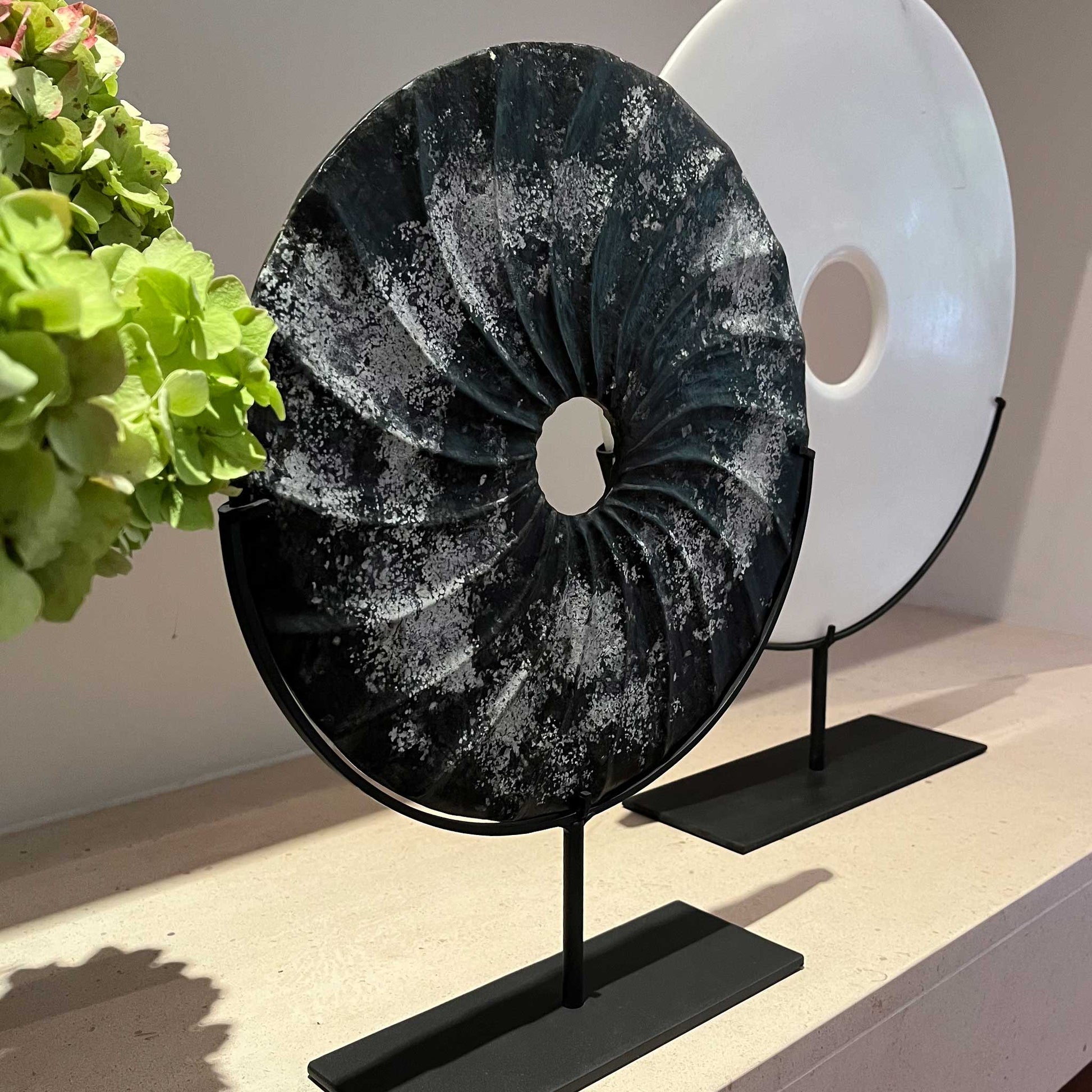 Lifestyle image of distressed black marble bi disc with shell type grooves displayed on a bronze stand. Next to larger plain white marble disc, also on a marble stand.