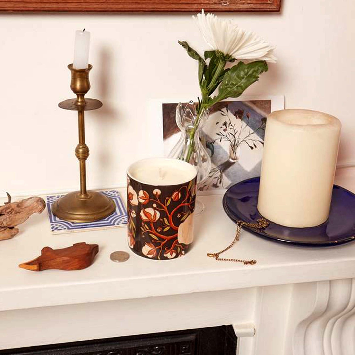 Lifestyle image of ceramic candle jar illustrated with open hands and cotton plants on mantelpiece with vintage pieces